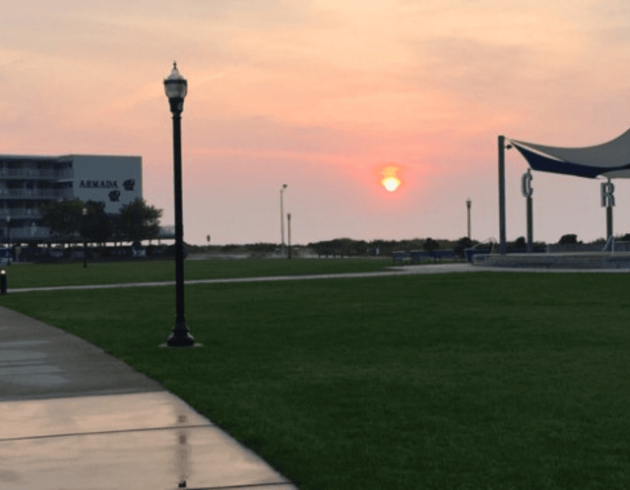 A sunset glows softly behind a park with green grass and a lamppost, accompanied by a modern building in the background.