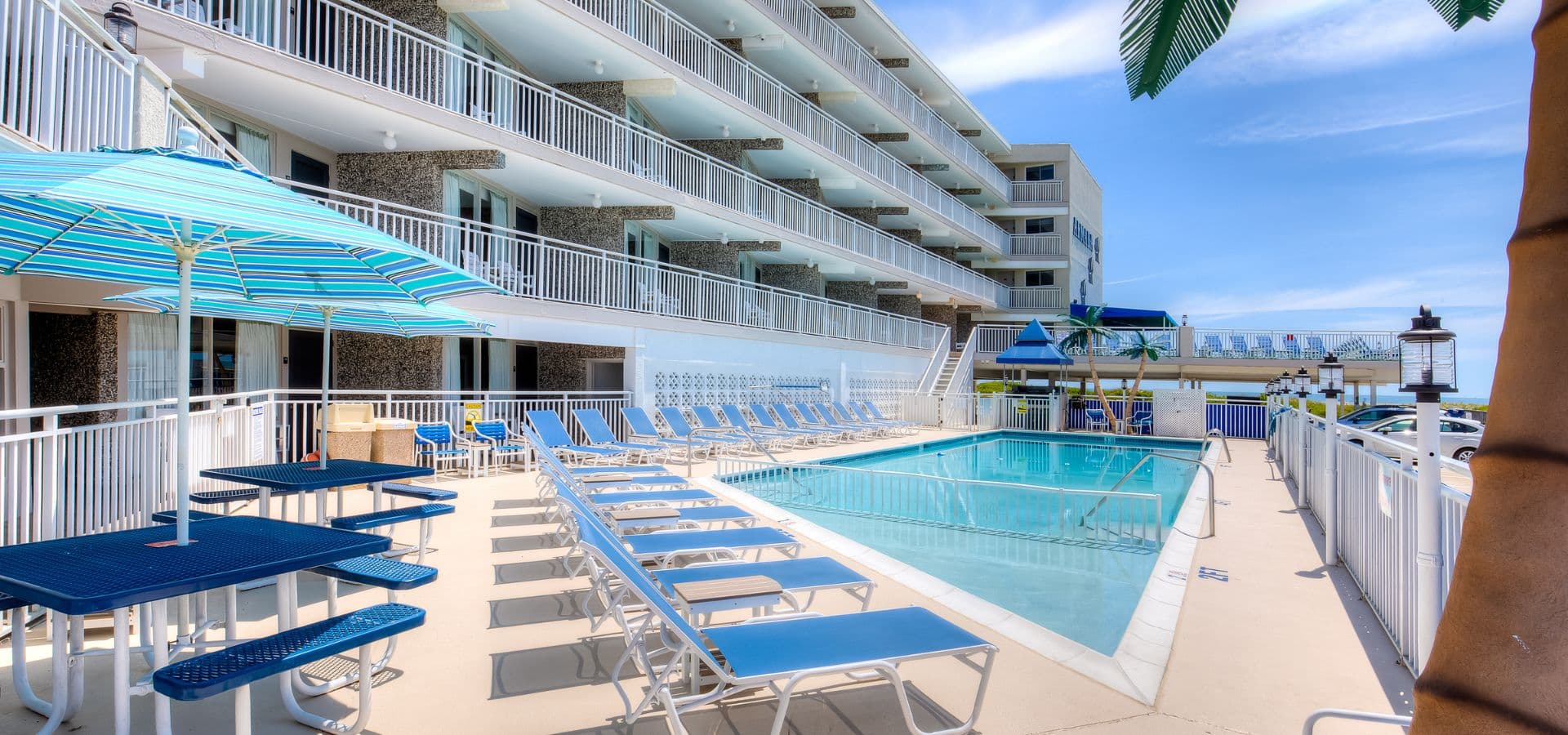 A sunny pool area with blue umbrellas and lounge chairs beside a multi-story hotel.