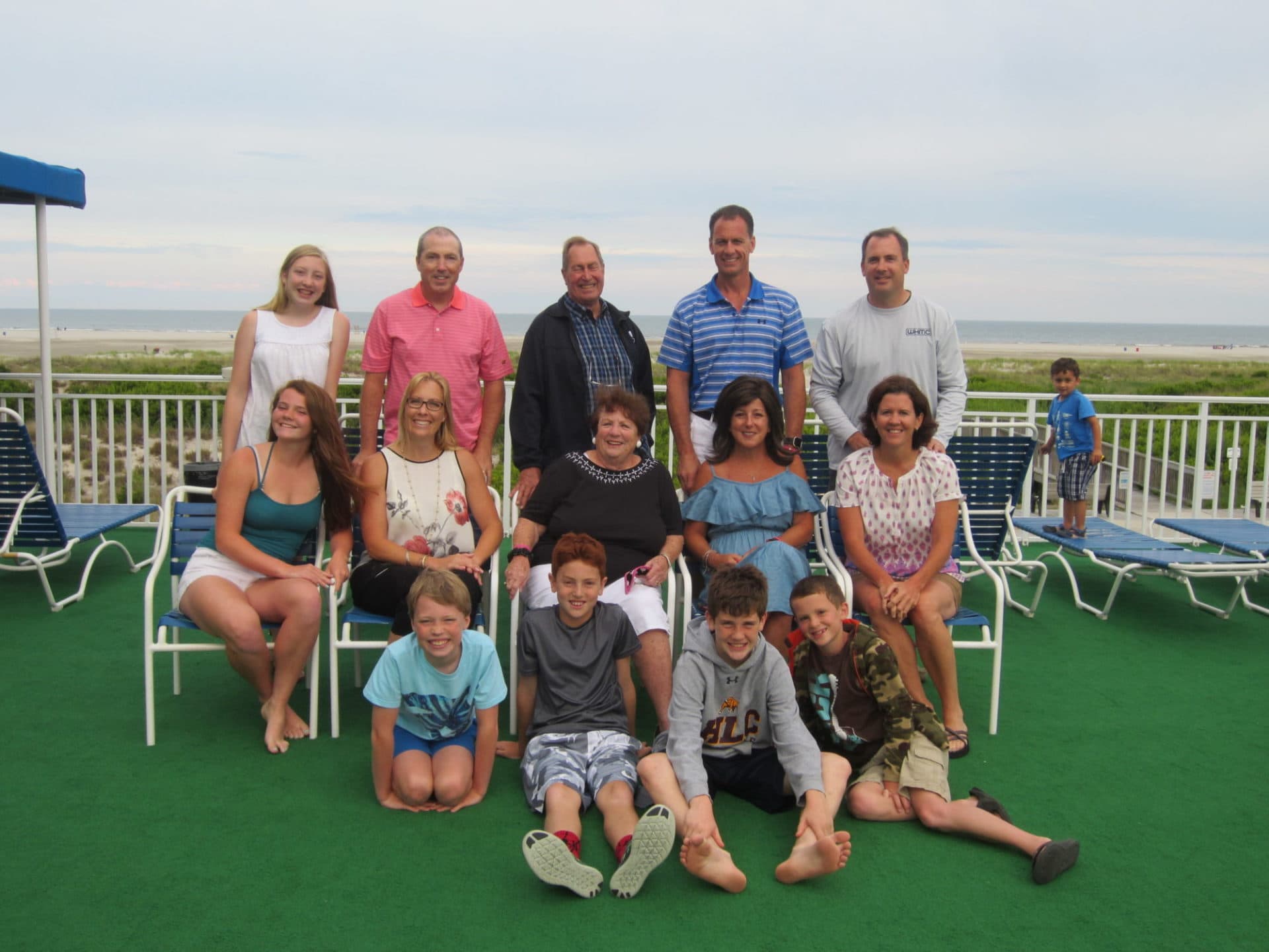 A large group of family members poses together on a deck with beach chairs and an ocean view in the background.
