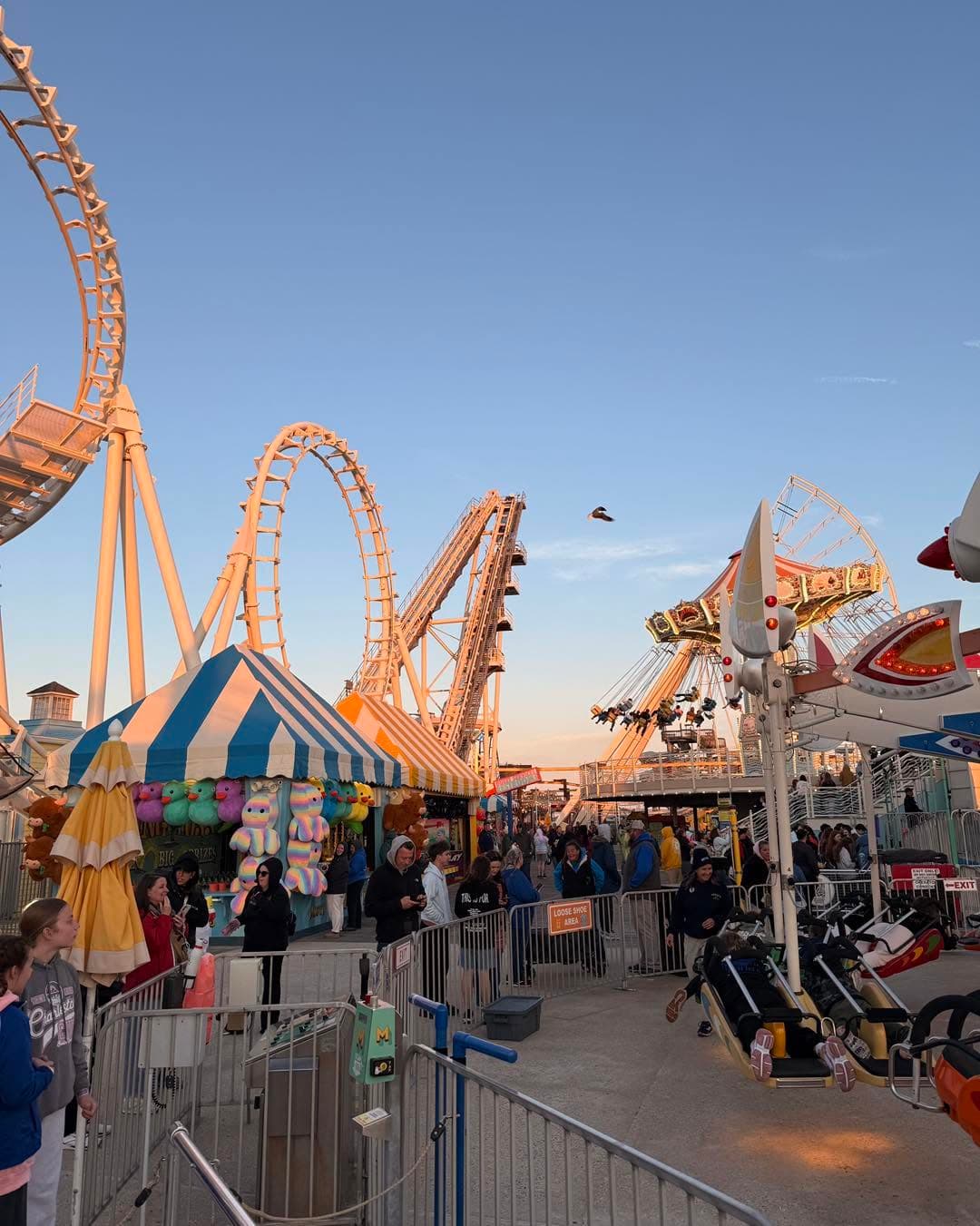 A vibrant amusement park scene featuring roller coasters, colorful game stalls, and a crowd of visitors enjoying the attractions at sunset.