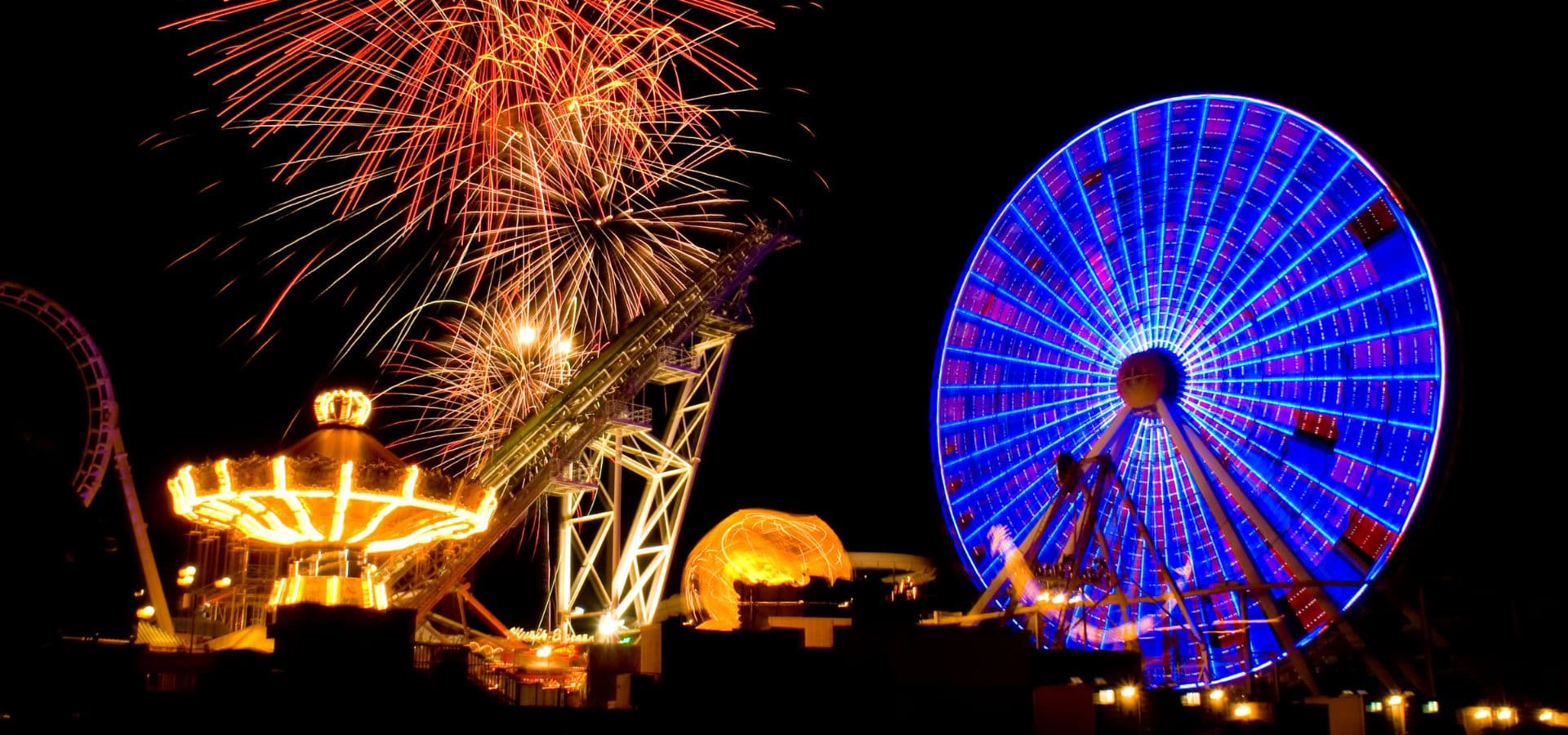 Colorful fireworks illuminate a nighttime amusement park featuring a brightly lit ferris wheel and carousel.
