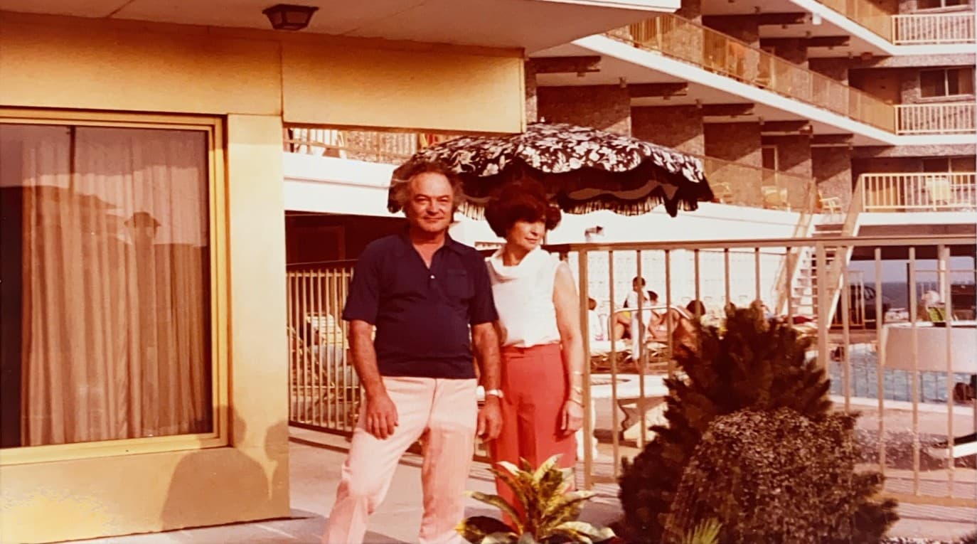 A man and woman stand together in front of a hotel balcony, with an umbrella and pool visible in the background.