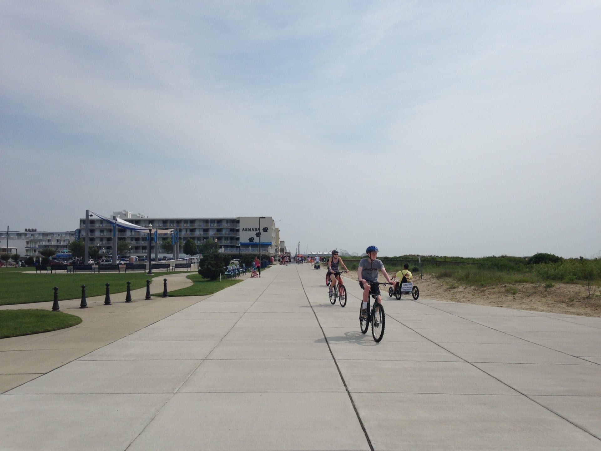 Bicyclists riding along a paved path with a beach and hotel in the background.