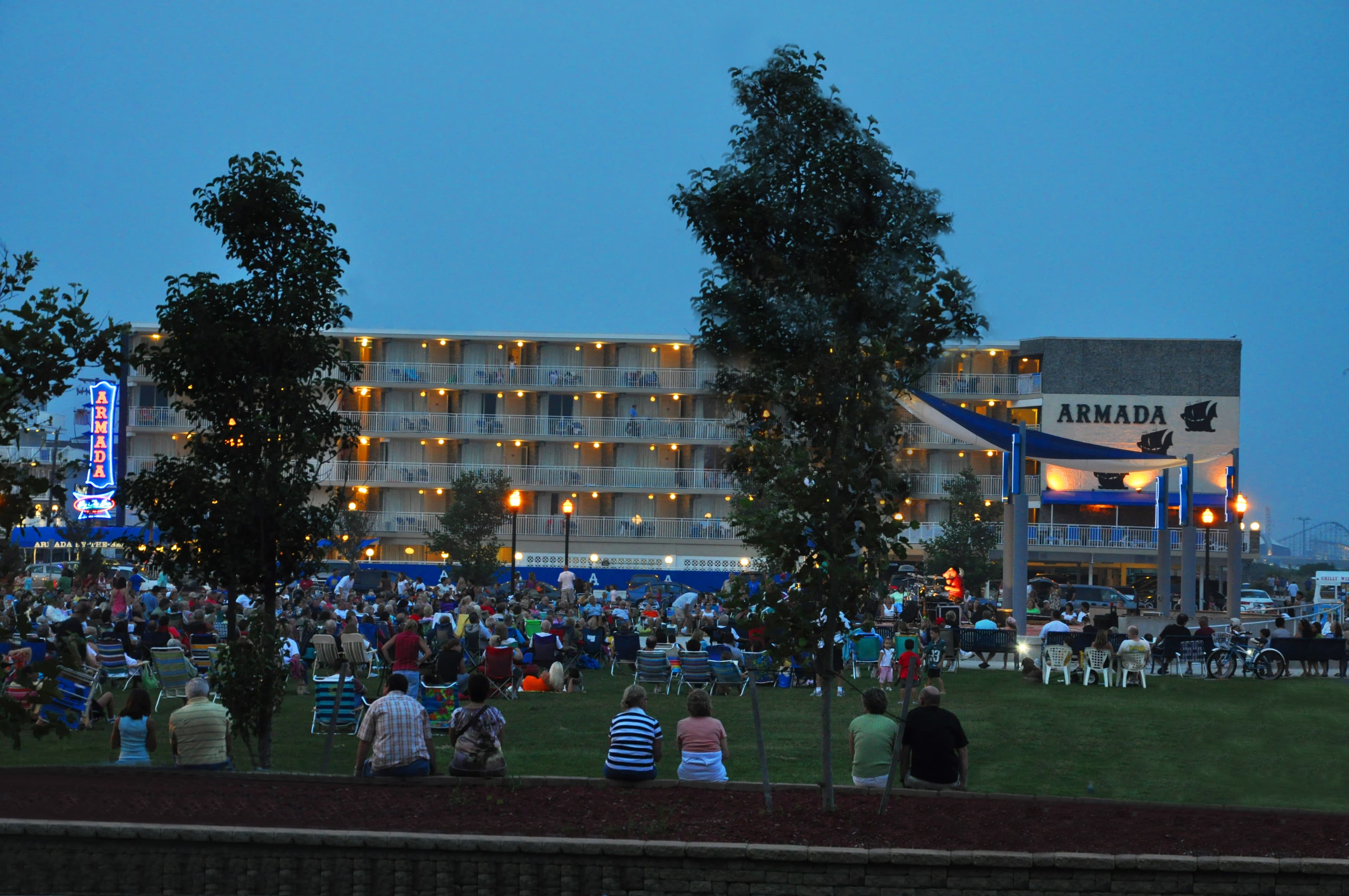 A crowd gathers on a lawn in front of a lit hotel named "Armada" during dusk.