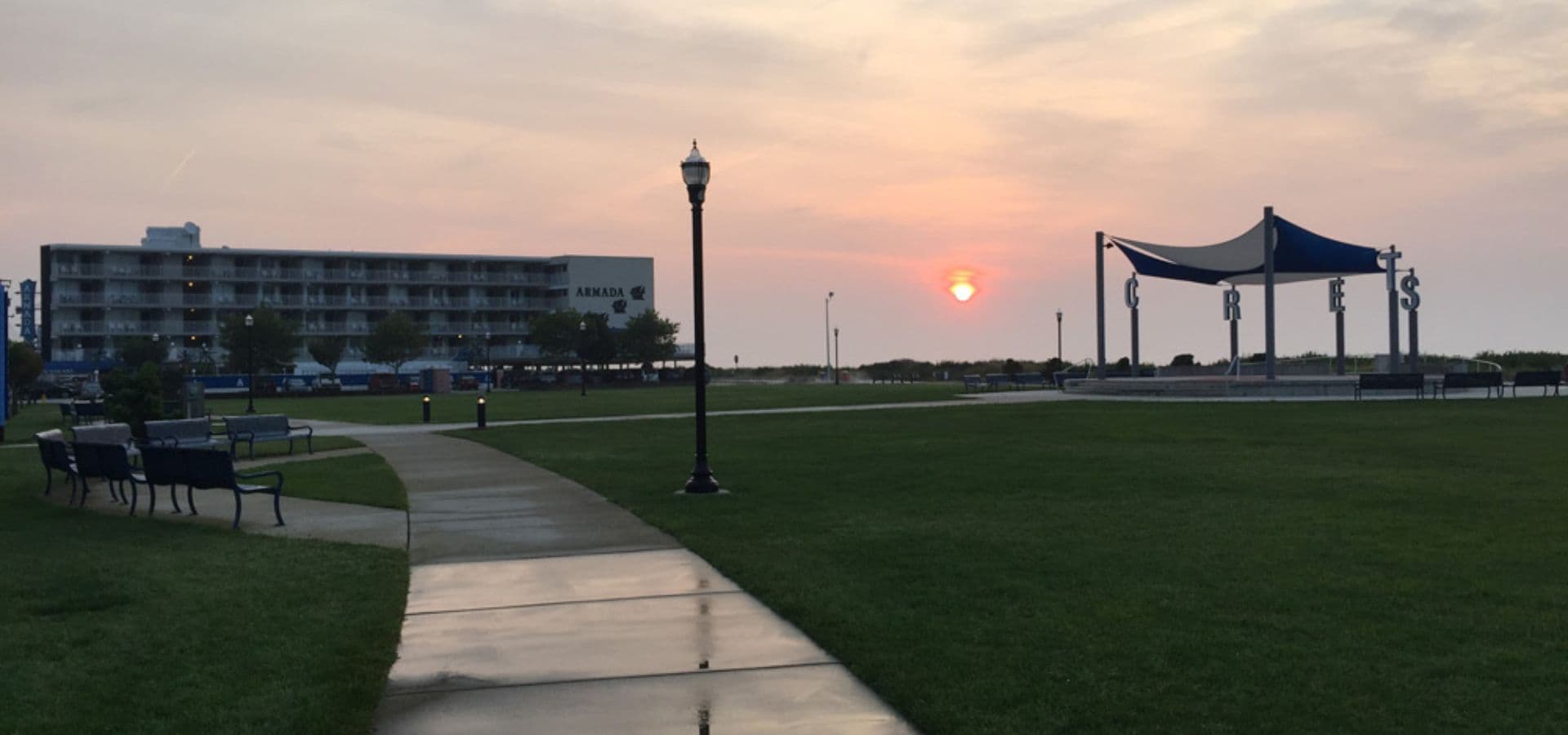A path leads through a grassy area towards a sunset, with a building and a pavilion in the background.