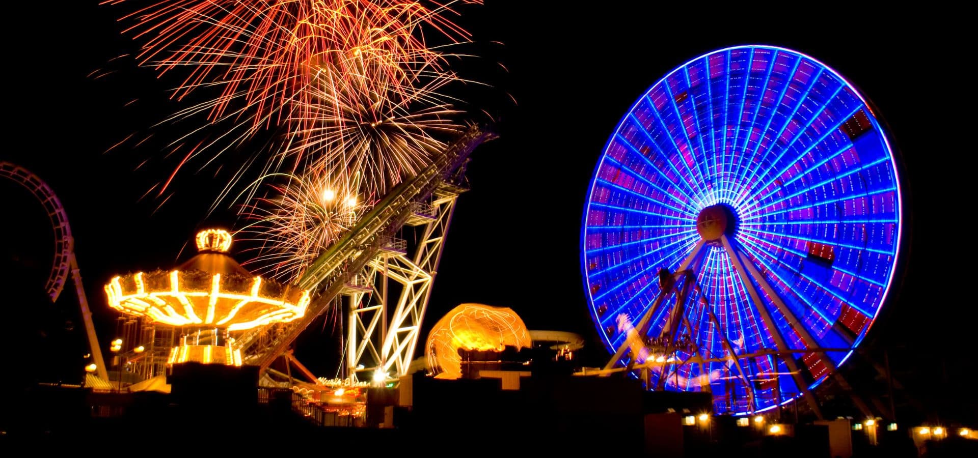 Colorful fireworks illuminate a night sky above a vibrant amusement park featuring a carousel and a large Ferris wheel.