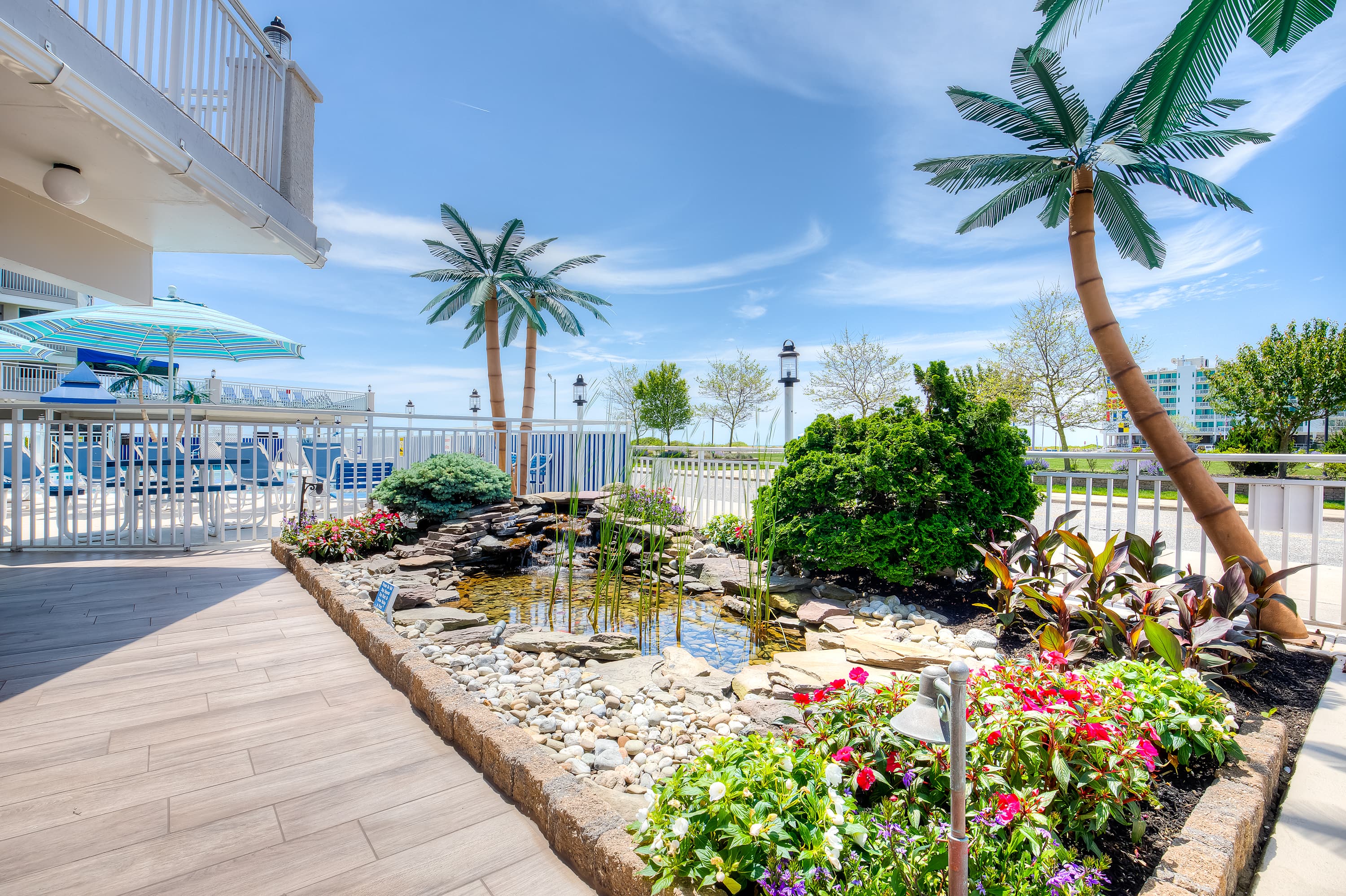 A serene outdoor area featuring a small pond surrounded by colorful flowers and tropical palm trees under a clear blue sky.