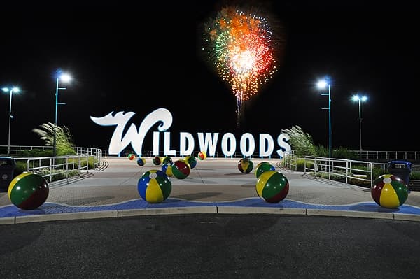 Fireworks illuminate the night sky above the "Wildwoods" sign surrounded by colorful beach balls.