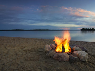 Camp fire on sandy beach, beside lake at sunset.