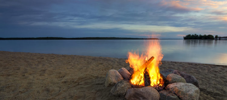 Camp fire on sandy beach, beside lake at sunset.